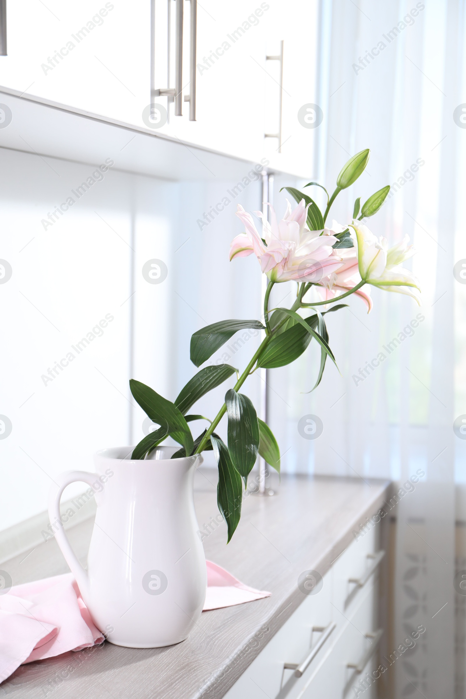 Beautiful lily flowers in jug on wooden cabinet indoors Photo of Beautiful lily flowers in jug on wooden cabinet indoors