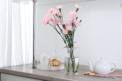 Photo of Beautiful carnation flowers in vase, cookies, teapot and cups on wooden cabinet indoors