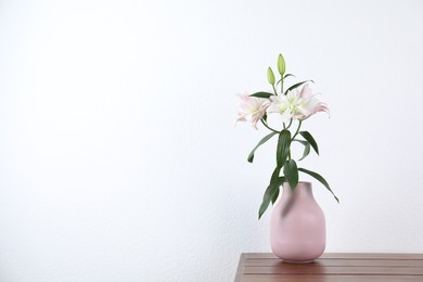 Photo of Beautiful lily flowers in vase on wooden table near white wall indoors, space for text