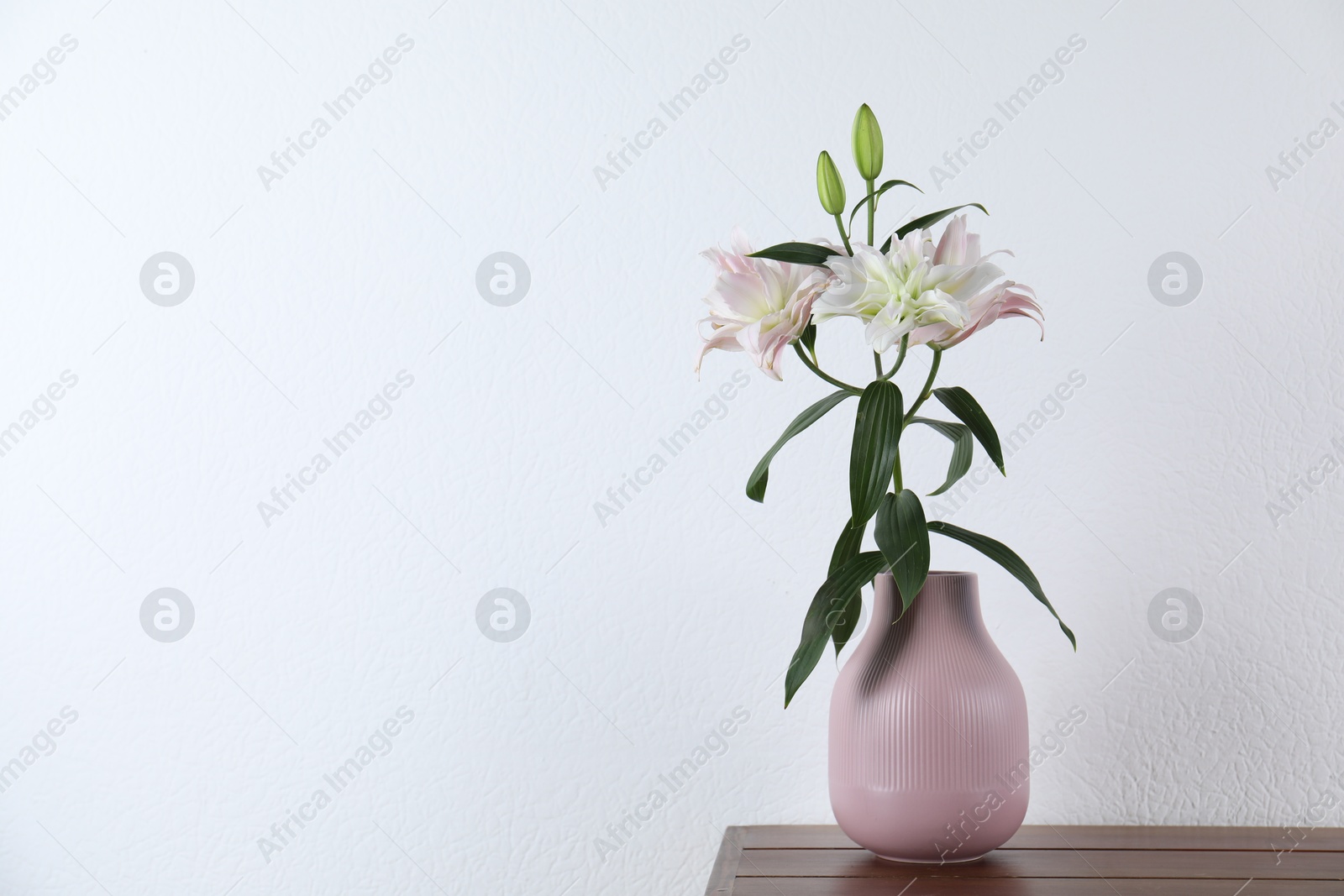 Photo of Beautiful lily flowers in vase on wooden table near white wall indoors, space for text