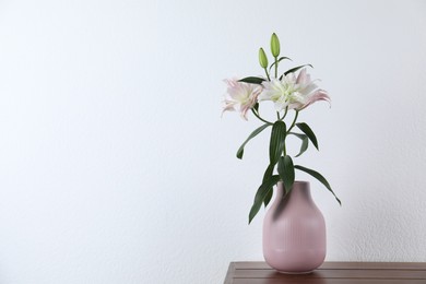 Photo of Beautiful lily flowers in vase on wooden table near white wall indoors, space for text