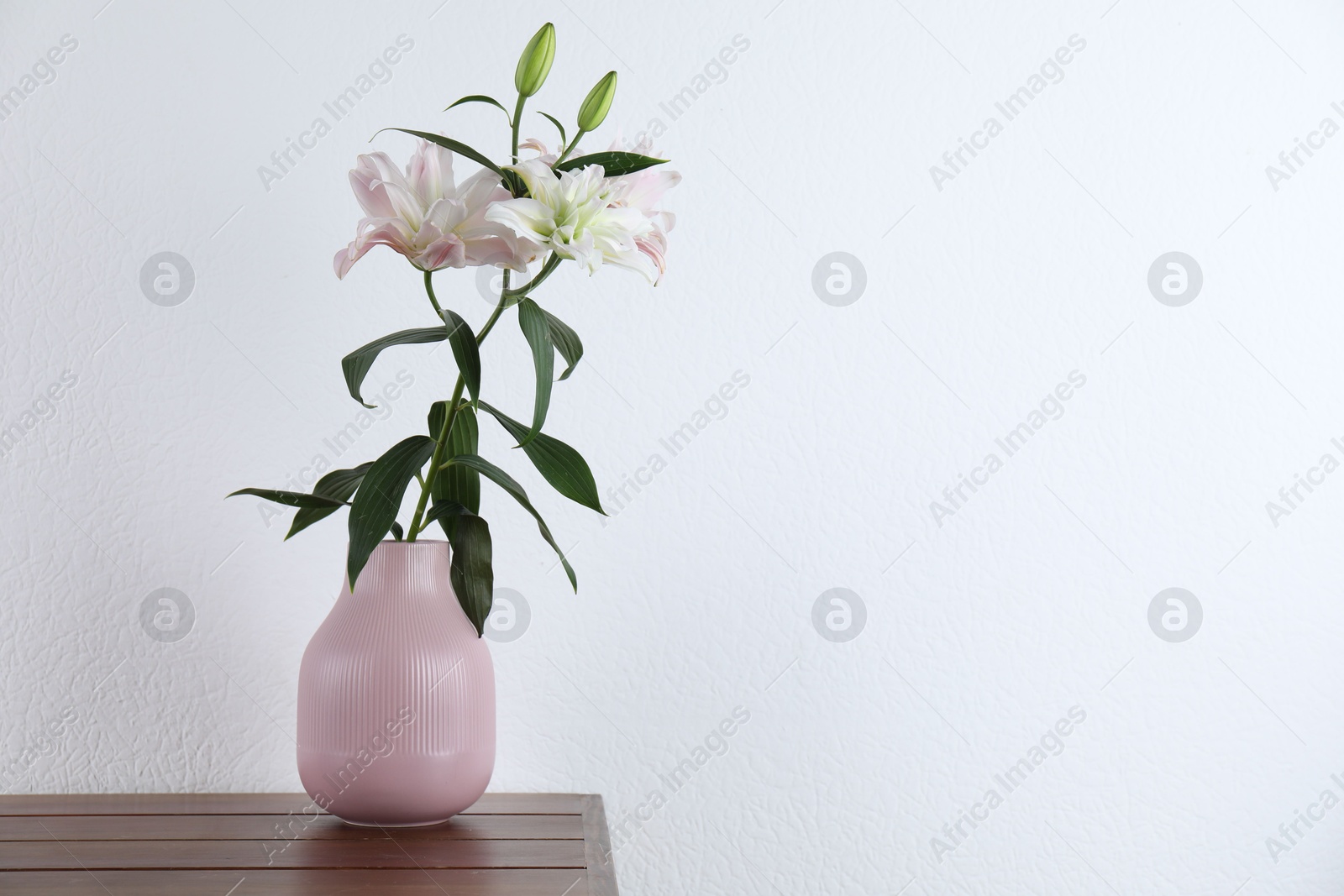 Beautiful lily flowers in vase on wooden table near white wall indoors, space for text Photo of Beautiful lily flowers in vase on wooden table near white wall indoors, space for text