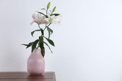 Photo of Beautiful lily flowers in vase on wooden table near white wall indoors, space for text
