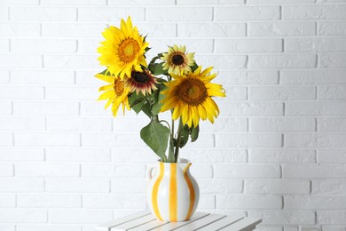 Photo of Beautiful sunflowers in jug on wooden table near white brick wall indoors