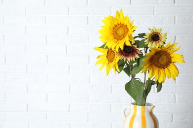 Photo of Beautiful sunflowers in jug against white brick wall indoors, space for text
