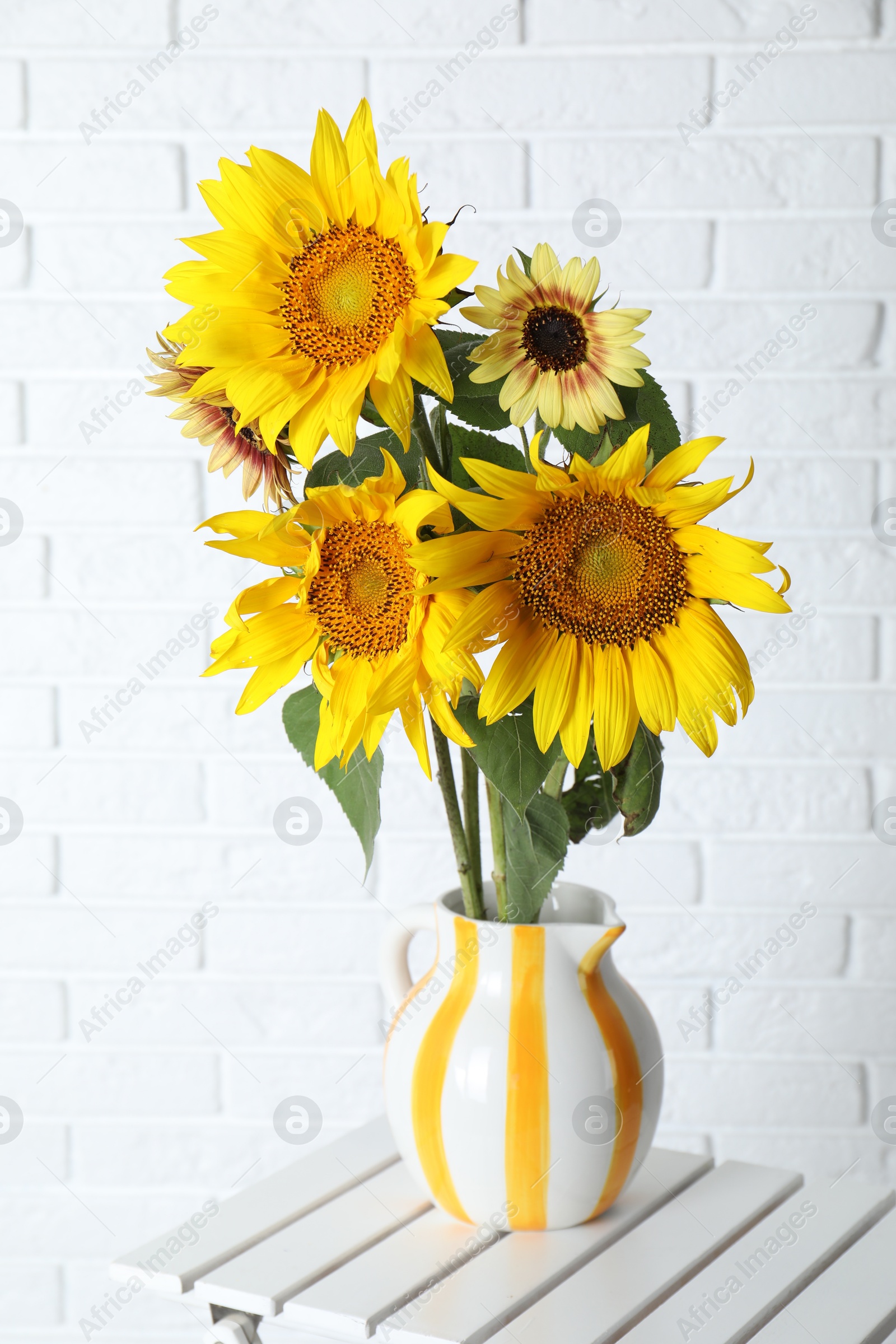 Photo of Beautiful sunflowers in jug on wooden table near white brick wall indoors