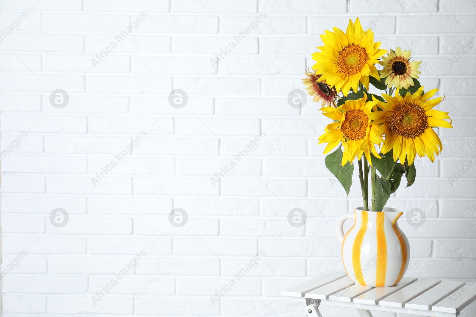 Photo of Beautiful sunflowers in jug on wooden table near white brick wall indoors, space for text