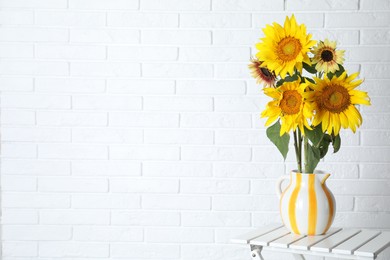Photo of Beautiful sunflowers in jug on wooden table near white brick wall indoors, space for text