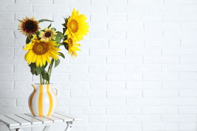 Photo of Beautiful sunflowers in jug on wooden table near white brick wall indoors, space for text