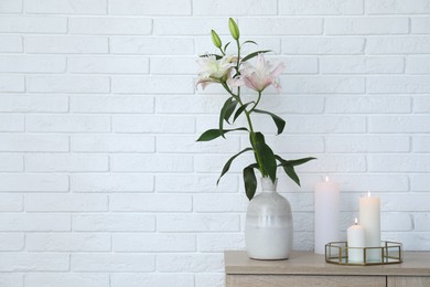 Photo of Beautiful lily flowers in vase and burning candles on cabinet near white brick wall indoors, space for text