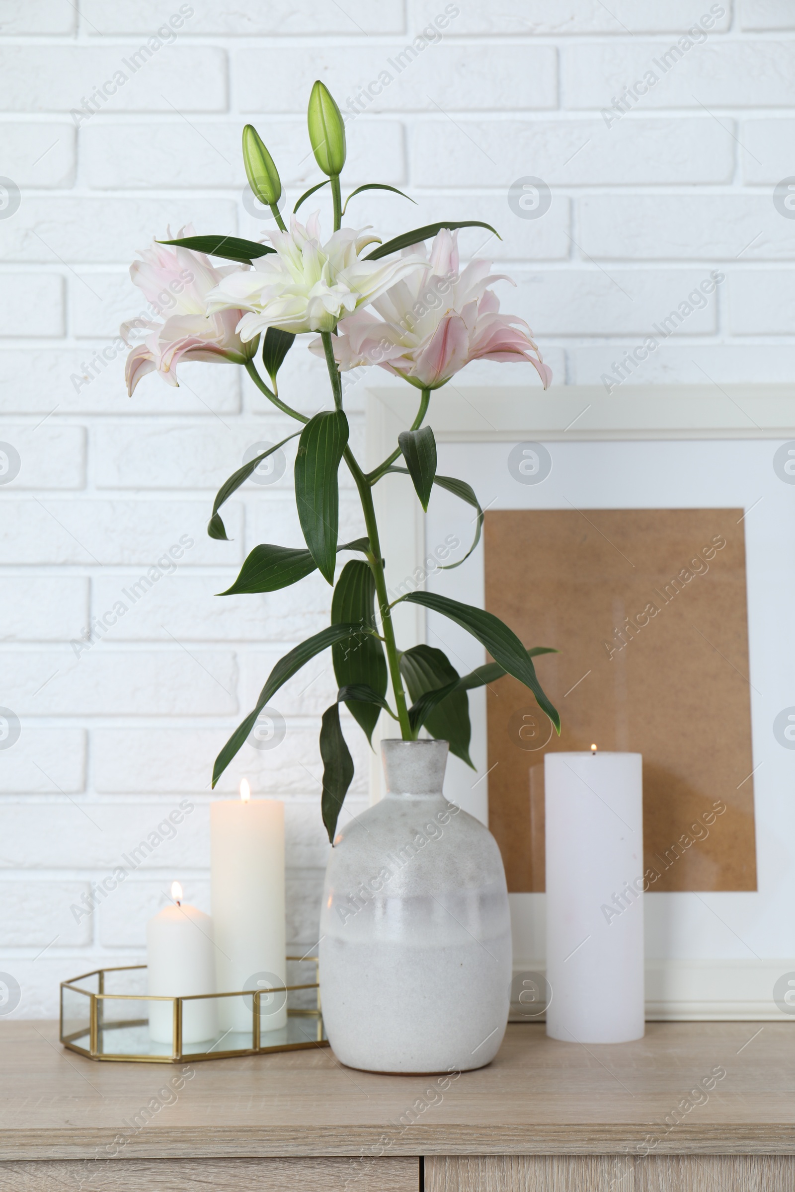 Photo of Beautiful lily flowers in vase and burning candles on cabinet near white brick wall indoors