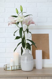 Photo of Beautiful lily flowers in vase and burning candles on cabinet near white brick wall indoors
