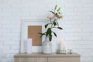 Photo of Beautiful lily flowers in vase and burning candles on cabinet near white brick wall indoors