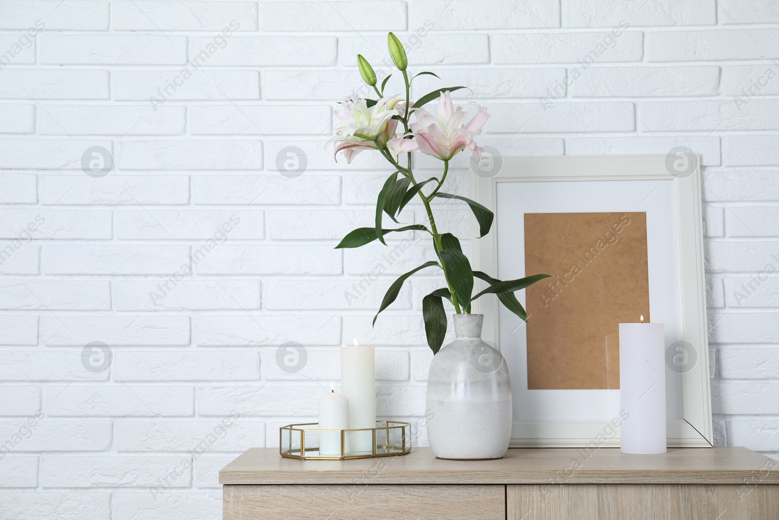 Beautiful lily flowers in vase and burning candles on cabinet near white brick wall indoors, space for text Photo of Beautiful lily flowers in vase and burning candles on cabinet near white brick wall indoors, space for text