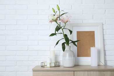 Photo of Beautiful lily flowers in vase and burning candles on cabinet near white brick wall indoors, space for text