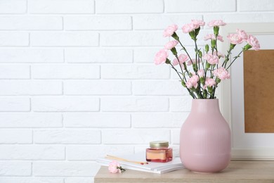 Photo of Beautiful carnation flowers in vase and other decor on wooden cabinet near white brick wall indoors, space for text