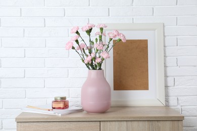 Photo of Beautiful carnation flowers in vase and other decor on wooden cabinet near white brick wall indoors