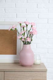 Photo of Beautiful carnation flowers in vase and other decor on wooden cabinet near white brick wall indoors