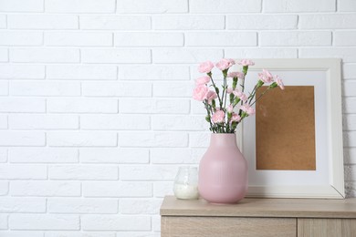 Photo of Beautiful carnation flowers in vase and other decor on wooden cabinet near white brick wall indoors, space for text