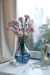 Photo of Beautiful carnation flowers in vase, mirror, jewellery and face roller near window indoors
