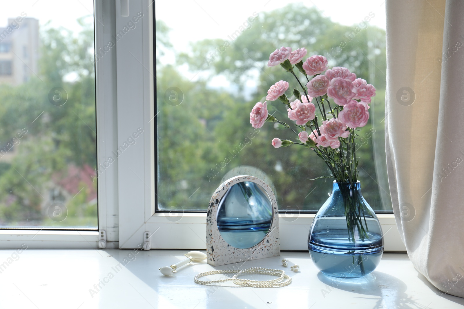 Beautiful carnation flowers in vase, mirror, jewellery and face roller near window indoors Photo of Beautiful carnation flowers in vase, mirror, jewellery and face roller near window indoors