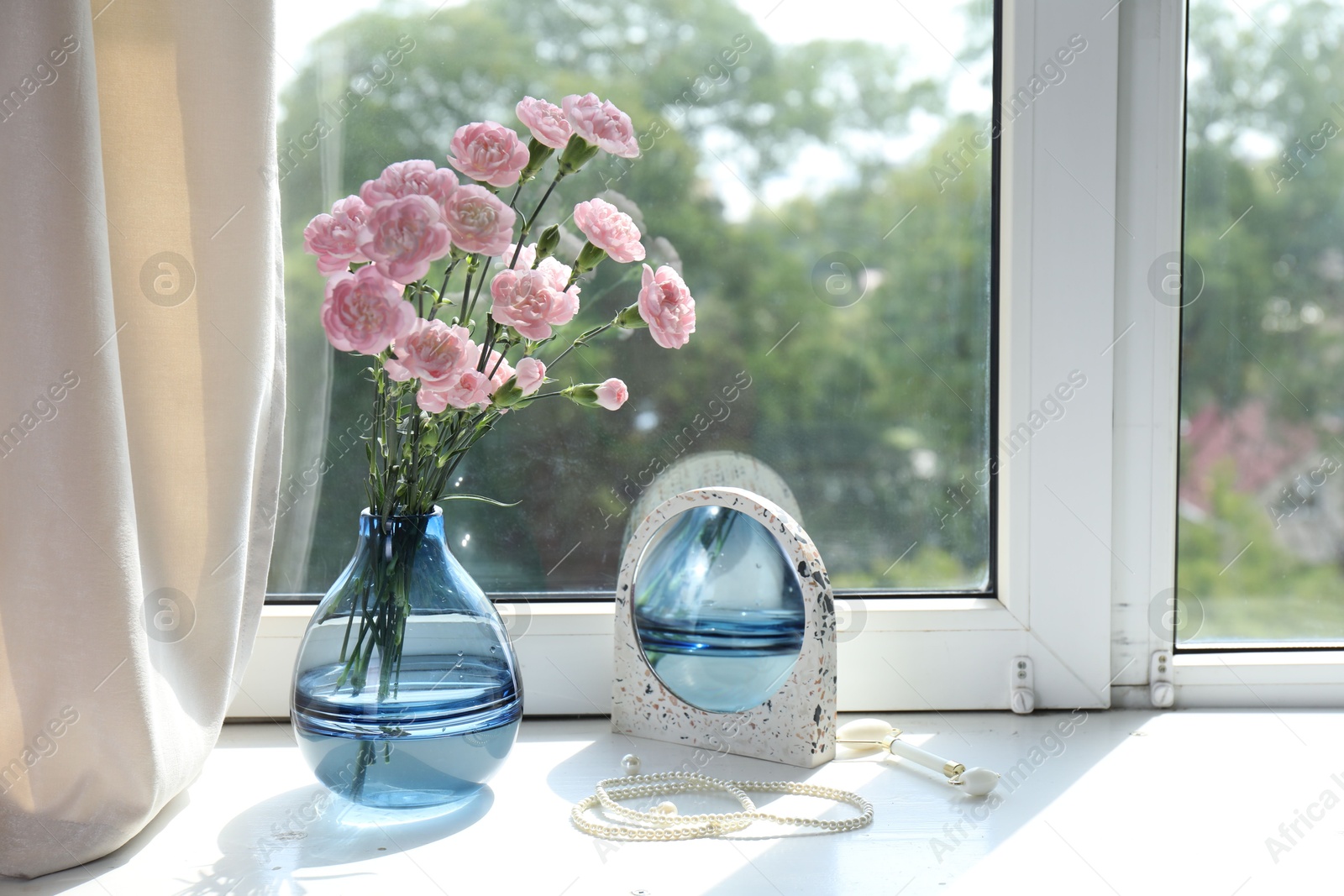 Photo of Beautiful carnation flowers in vase, mirror, jewellery and face roller near window indoors