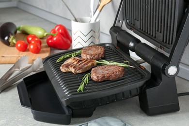 Photo of Open electric grill with beef steak, cutlets and rosemary on light grey table, closeup