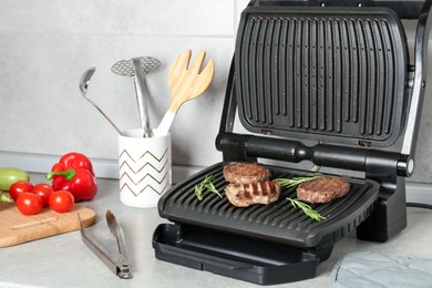 Photo of Open electric grill with beef steak, cutlets, rosemary, vegetables and cookware on light grey table, closeup