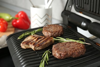 Photo of Open electric grill with beef steak, cutlets and rosemary on table, closeup