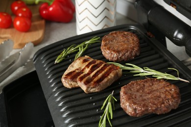 Photo of Open electric grill with beef steak, cutlets and rosemary on table, closeup