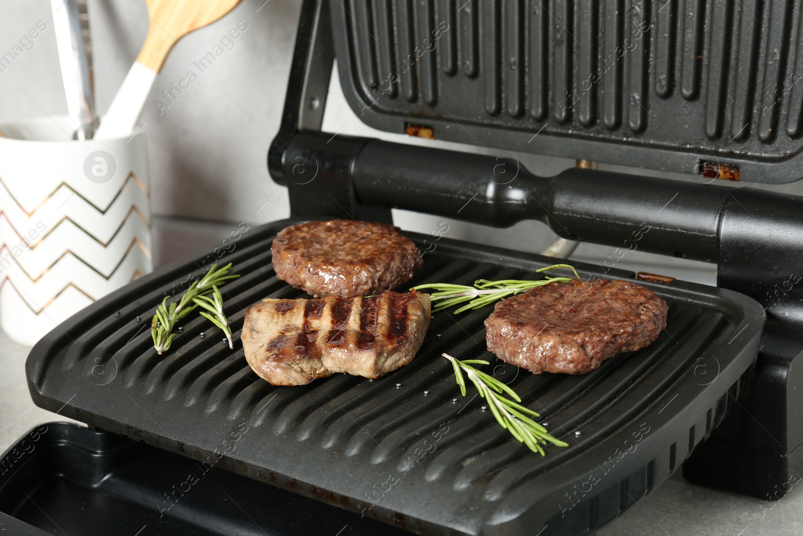 Photo of Open electric grill with beef steak, cutlets and rosemary on grey table, closeup