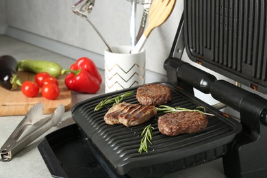Photo of Open electric grill with beef steak, cutlets and rosemary, vegetables and cookware on light grey table, closeup