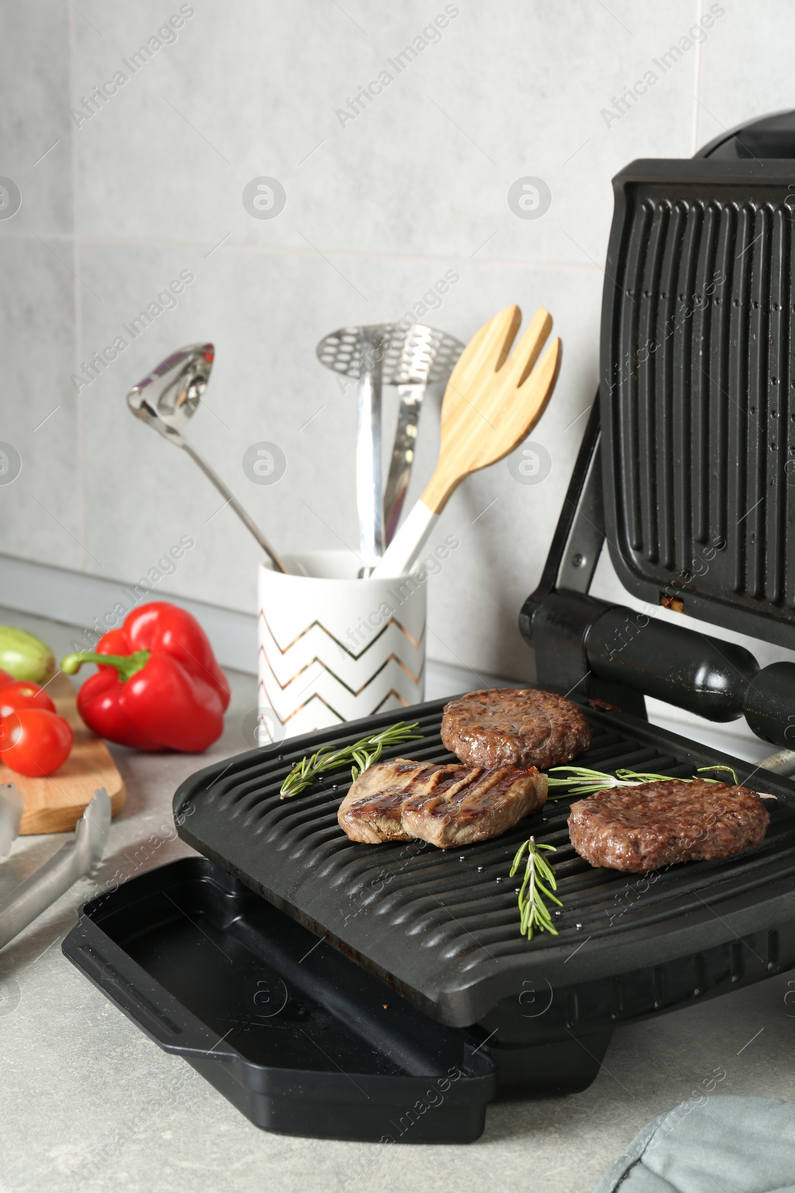 Photo of Open electric grill with beef steak, cutlets and rosemary, vegetables and cookware on light grey table, closeup