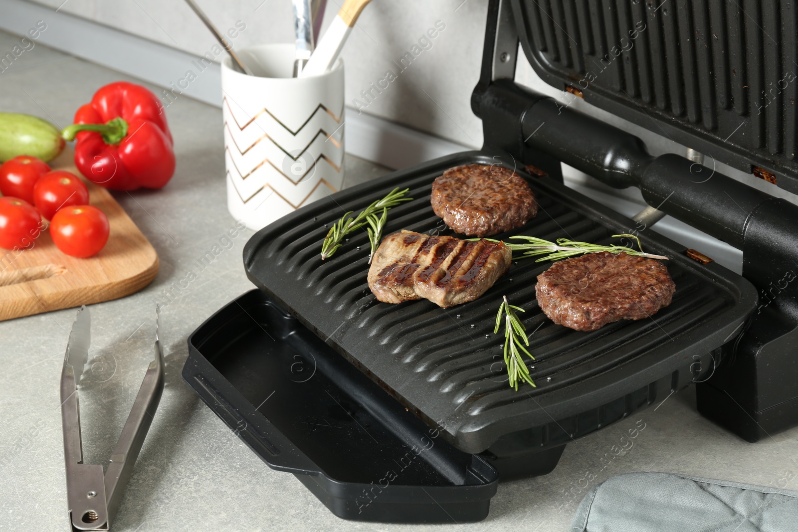 Photo of Open electric grill with beef steak, cutlets and rosemary, vegetables and cookware on light grey table, closeup