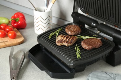 Photo of Open electric grill with beef steak, cutlets and rosemary, vegetables and cookware on light grey table, closeup