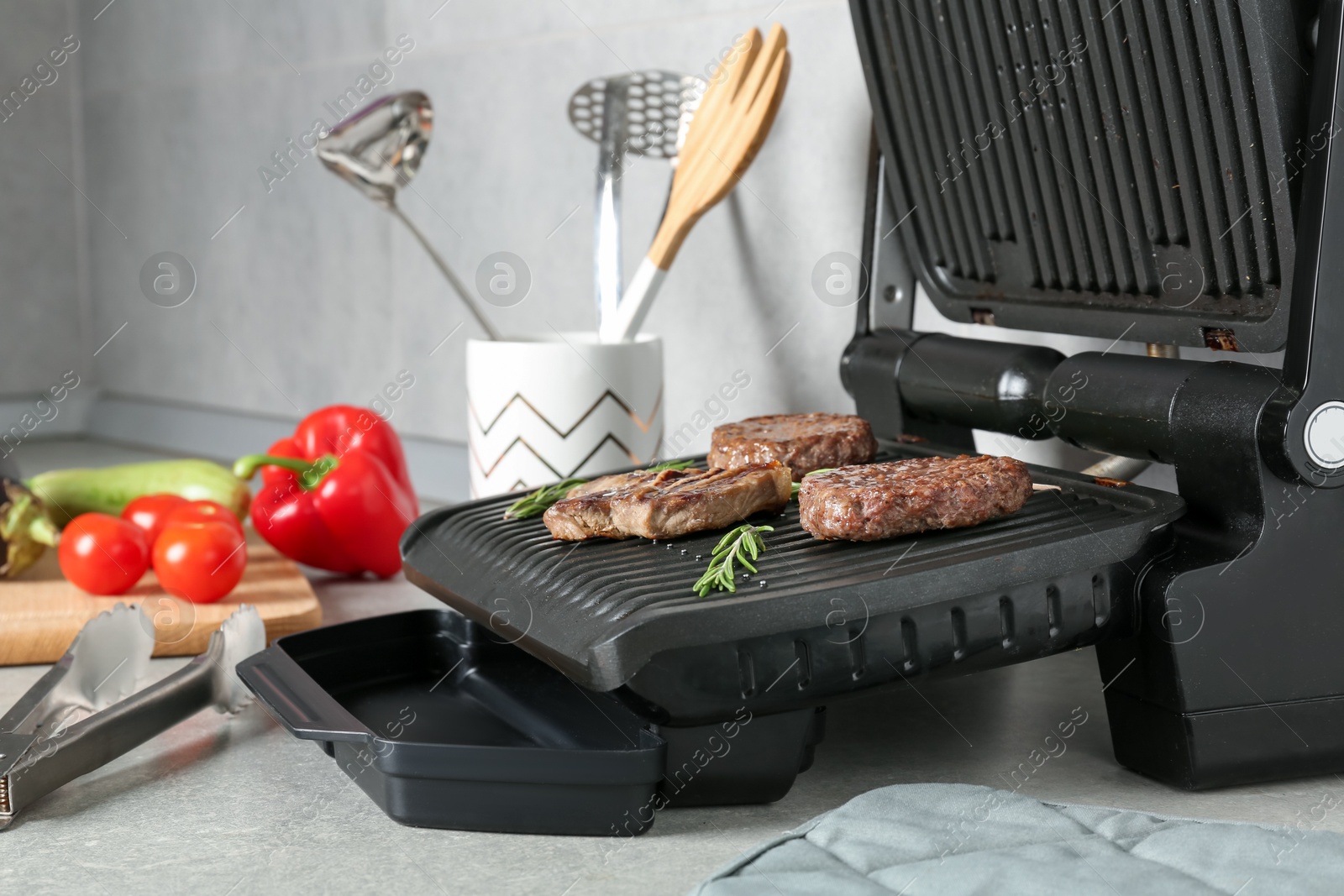 Photo of Open electric grill with beef steak, cutlets and rosemary, vegetables and cookware on light grey table, closeup