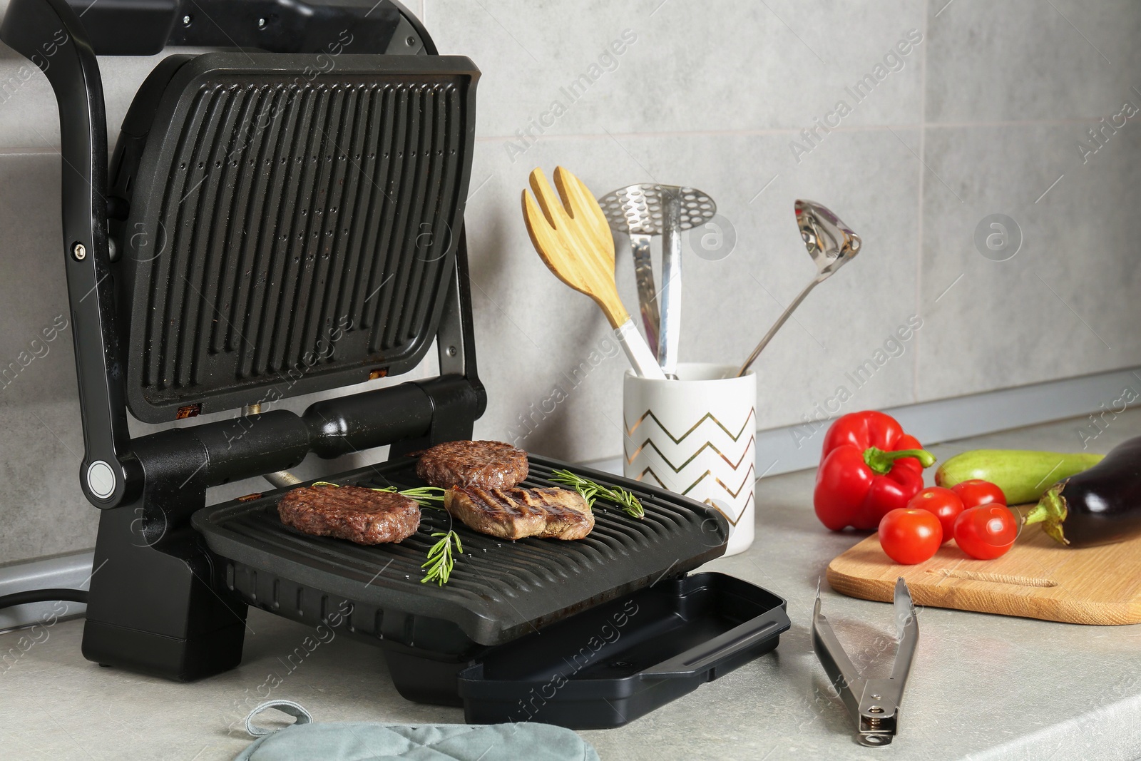 Photo of Open electric grill with beef steak, cutlets and rosemary, vegetables and cookware on light grey table, closeup