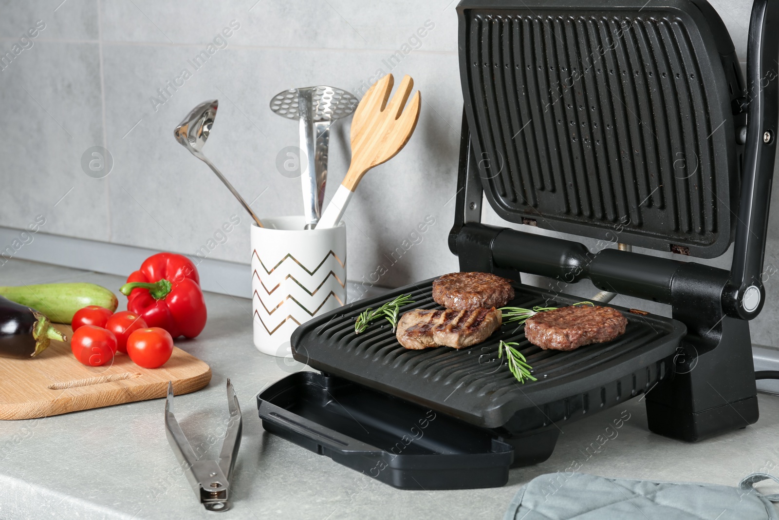 Photo of Open electric grill with beef steak, cutlets and rosemary, vegetables and cookware on light grey table, closeup