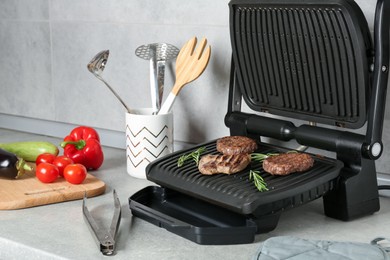 Photo of Open electric grill with beef steak, cutlets and rosemary, vegetables and cookware on light grey table, closeup