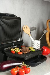 Photo of Open electric grill with beef steak, zucchini, tomatoes and cookware on light grey table