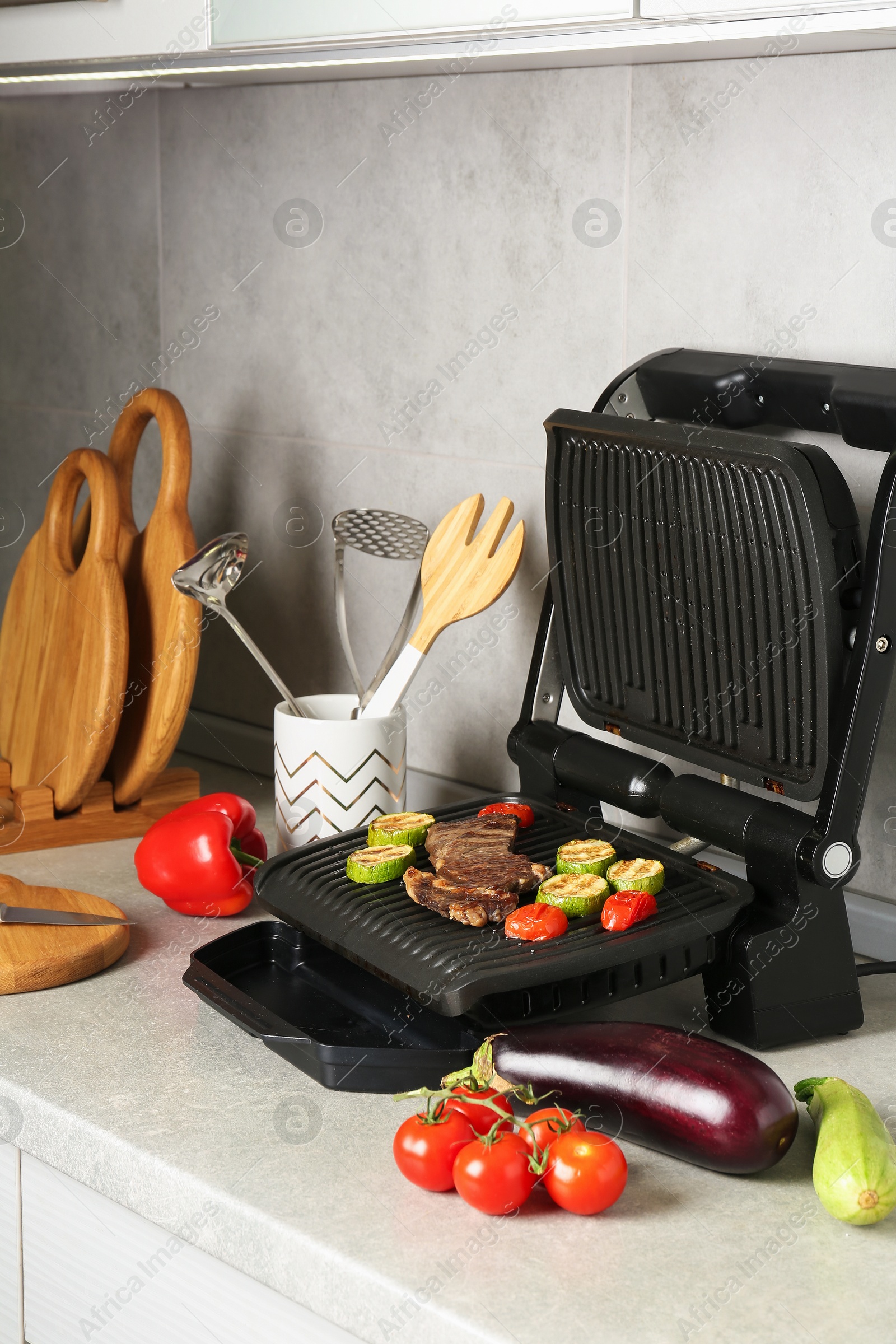 Open electric grill with beef steak, zucchini, tomatoes and cookware on light grey table Photo of Open electric grill with beef steak, zucchini, tomatoes and cookware on light grey table