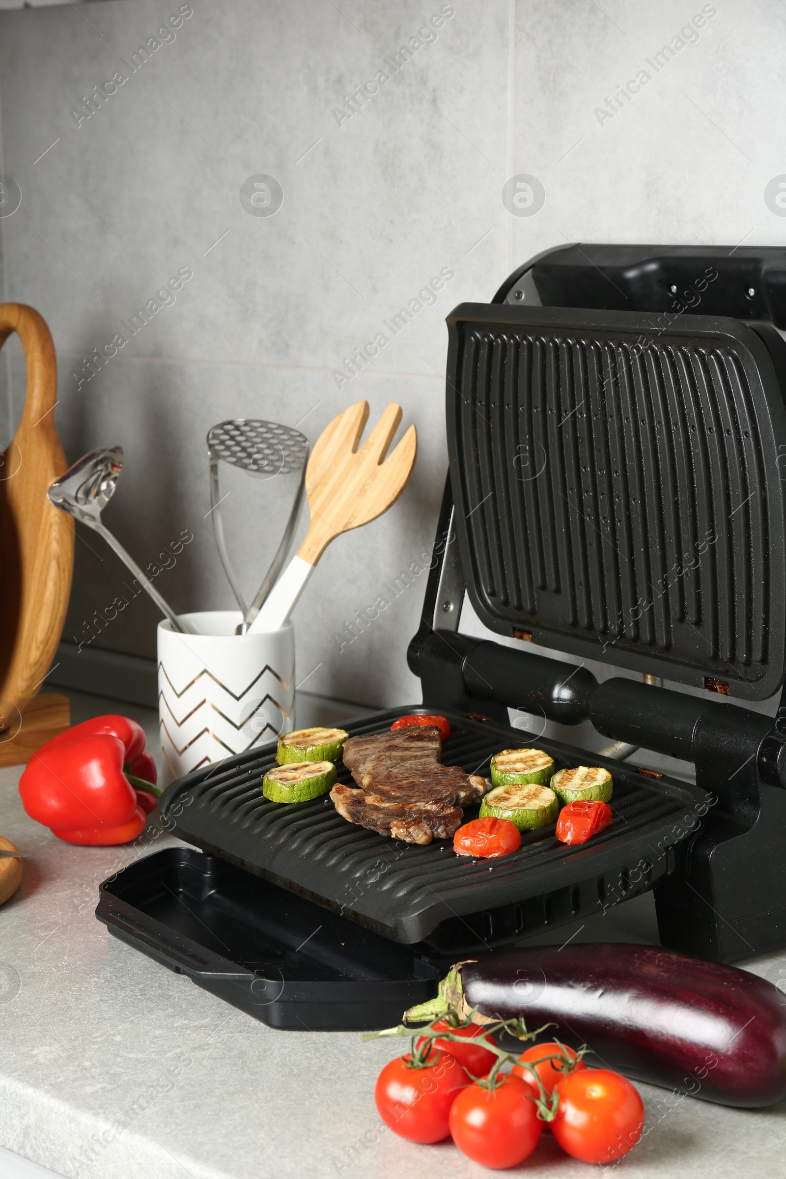Open electric grill with beef steak, zucchini, tomatoes and cookware on light grey table Photo of Open electric grill with beef steak, zucchini, tomatoes and cookware on light grey table