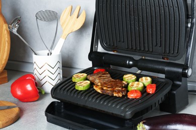 Photo of Open electric grill with beef steak, zucchini, tomatoes and cookware on light grey table, closeup
