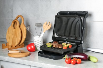 Photo of Open electric grill with beef steak, vegetables and cookware on light grey table