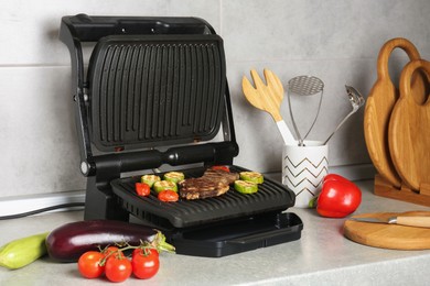 Photo of Open electric grill with beef steak, vegetables and cookware on light grey table