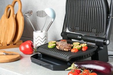Photo of Open electric grill with beef steak, vegetables and cookware on light grey table, closeup