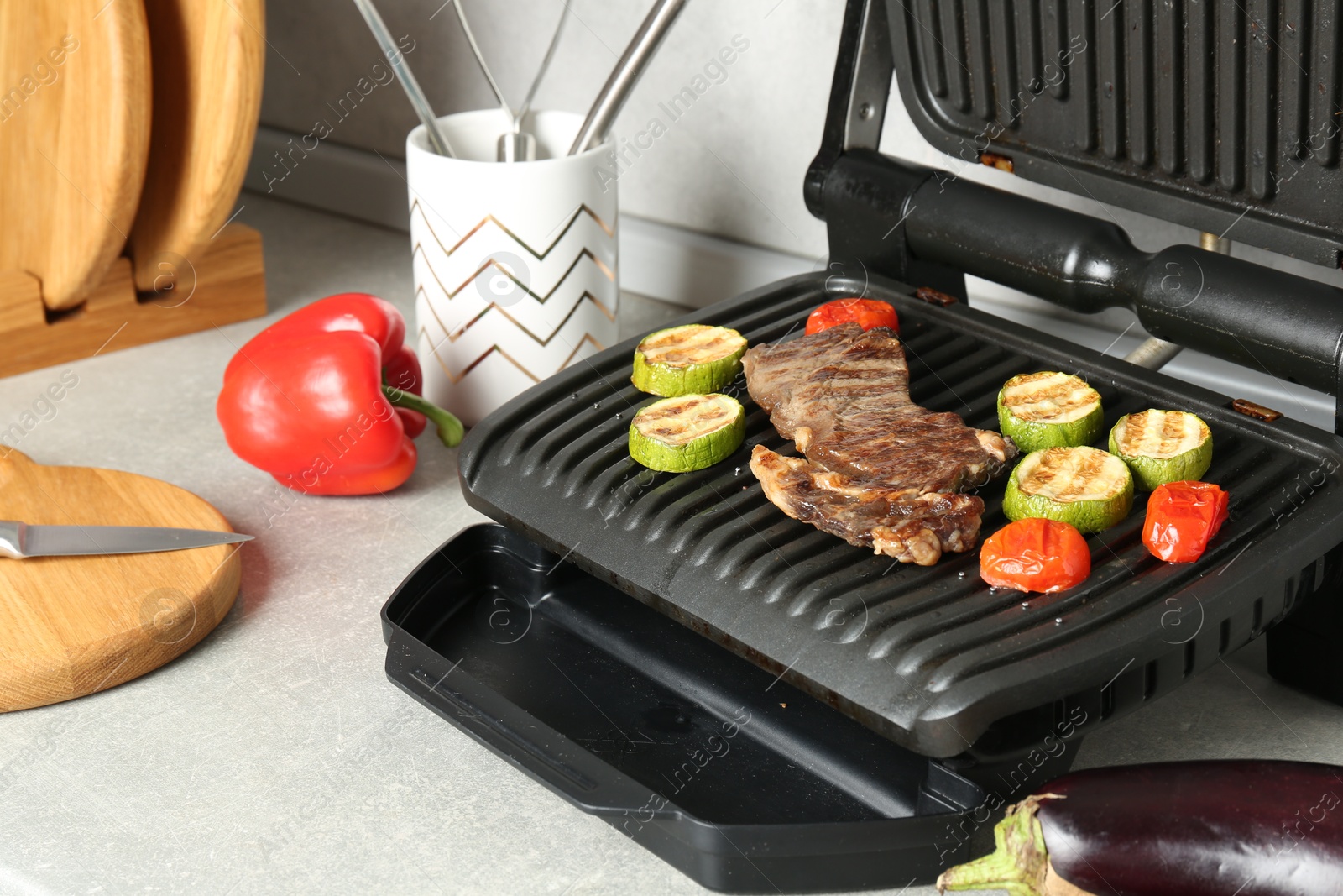 Photo of Open electric grill with beef steak and vegetables on light grey table, closeup