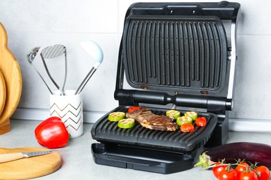 Photo of Open electric grill with beef steak, vegetables and cookware on light grey table