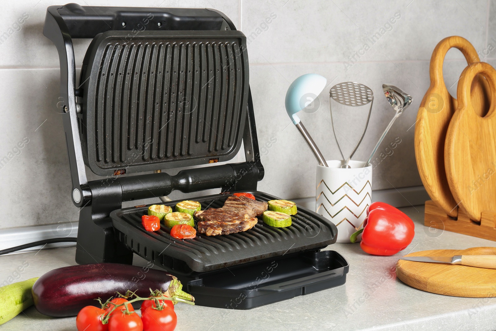Photo of Open electric grill with beef steak, vegetables and cookware on light grey table