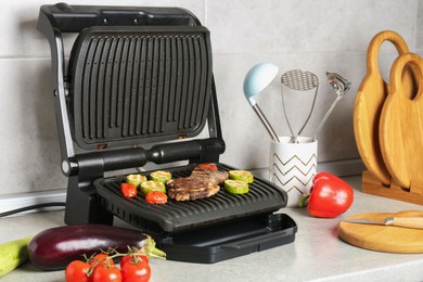 Photo of Open electric grill with beef steak, vegetables and cookware on light grey table